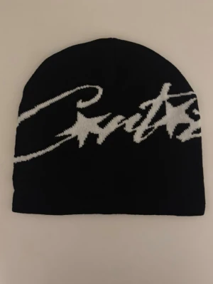 Svart mössa med vit text  - Snygg svart mössa  med vit, stor logotyp i graffiti-stil över hela framsidan. Klassisk passform och mjukt stickat material som håller dig varm. Perfekt accessoar för en streetwear-look.