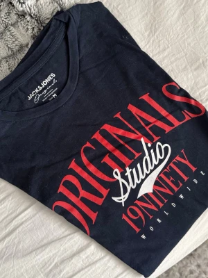 Svart t-shirt Jack & Jones Originals - Snygg svart t-shirt från Jack & Jones Originals med röd och vit tryckt text på bröstet. Modellen har rund halsringning och är tillverkad i mjuk bomull. Perfekt för en avslappnad och trendig look.