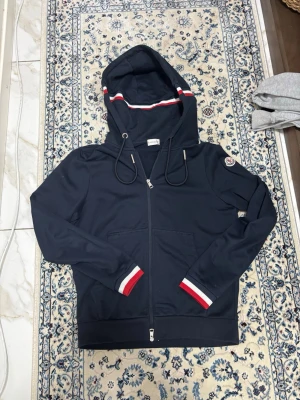 Marinblå hoodie från Moncler - Snygg marinblå hoodie från Moncler med dragkedja, huva och vita samt röda detaljer på muddar och huva. Klassisk Moncler-logga på ärmen och praktiska fickor framtill. Perfekt för en stilren och sportig look.