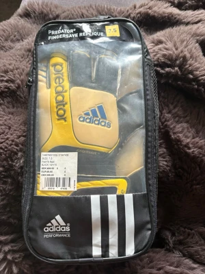Adidas Predator målvaktshandskar 7.5 - Säljer ett par Adidas Predator Fingersave Replique målvaktshandskar i svart och gult med blå logga. Handskarna har Fingersave-skydd för extra stöd och grepp, samt elastisk handledsrem för bästa passform. Perfekta för fotbollsmatcher och träning. Använd fåtal gånger - lite sliten se bild på ena handsken annars bra skick