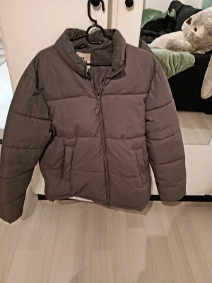 Grå pufferjacka med dragkedja - Snygg grå pufferjacka med hög krage och dragkedja framtill. Jackan har två stora fickor på sidorna och quiltad design som ger en cool och modern look. Perfekt för kalla vinterdagar när du vill hålla dig varm och stilren.