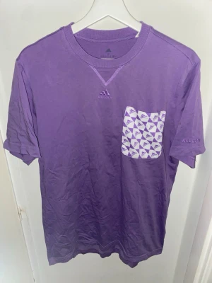 Lila Adidas t-shirt med ficka - Säljer en lila t-shirt från Adidas med en vit och lila mönstrad bröstficka. T-shirten har korta ärmar, rund halsringning och Adidas-logga broderad på bröstet. Tillverkad i mjuk bomull, perfekt för en avslappnad stil. Storlek S