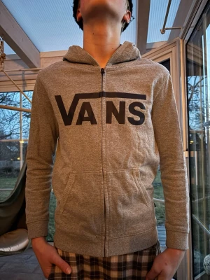 Grå zip-hoodie från Vans - Grå hoodie med dragkedja från Vans, stor svart logga framtill. Mjuk bomullsblandning, huva och två fickor. Perfekt för en avslappnad streetstil.