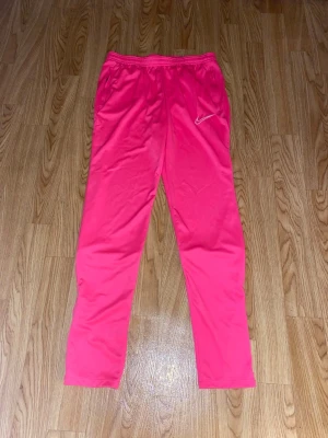 Nike DRI-FIT träningsbyxor  - Rosa träningsbyxor från Nike med smal passform och elastisk midja. Byxorna har en vit Nike-logga, två fickor med dragkedja och dragkedjor vid bensluten för enkel av- och påtagning. Storlek S, aldrig använda