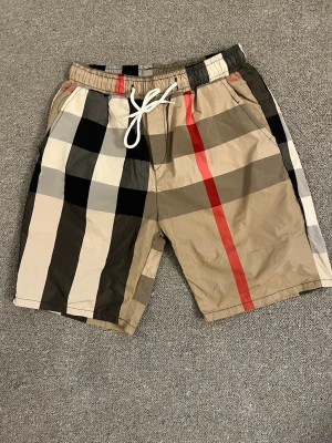 Rutig shorts med snörning Burberry - Säljer ett par klassiska rutiga shorts från Burberry med beige, svart, vit och röd färg. Shortsen har elastisk midja med vit snörning och sidofickor. Perfekta för sommaren och har den ikoniska Burberry-looken.