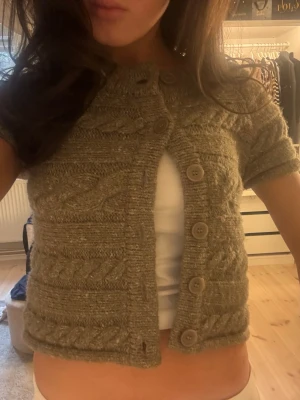 Kort stickad kofta med flätmönster - Säljer en beige, kort stickad kofta med flätmönster och stora knappar framtill. Koftan har korta ärmar och rund halsringning. Perfekt att styla över ett linne eller t-shirt för en trendig lager-på-lager-look.