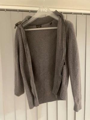 Grå kofta med huva från Massimo Dutti - Mysig grå kofta med dragkedja och vadderad huva. Koftan är ribbstickad och har långa ärmar, perfekt för lager-på-lager. Snygg och enkel att matcha till olika outfits.