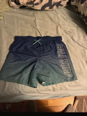 Blå badbyxor CALI SURF från H&M - Säljer ett par blå badbyxor från H&M med texten CALI SURF längs ena benet. De har resår i midjan och ett turkost snöre framtill. Materialet är snabbtorkande och insidan har meshfoder. Perfekta för stranden eller poolen!