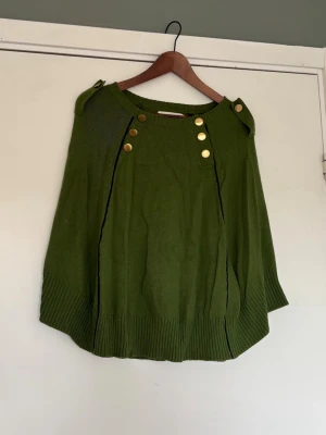 Grön stickad poncho med guldknappar - Cool grön stickad poncho med snygga guldknappar vid axlarna och svarta detaljer längs sömmarna. 