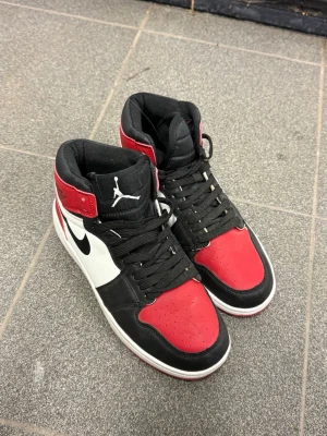 Air Jordan 1 Mid röd/svart/vit - Säljer ett par Air Jordan 1 Mid sneakers i klassisk röd, svart och vit färgkombination. Skorna har svart swoosh, vita paneler och röda detaljer på tå och häl. Hög komfort med vadderad krage och ikoniska Jordan-loggor på plösen och sidan. Perfekta för dig som gillar streetwear.