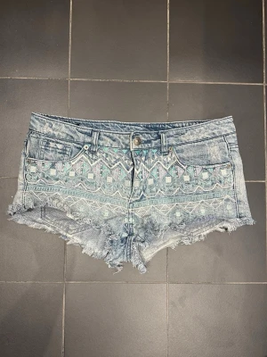 Blå jeansshorts med broderi - Säljer ett par ljusblå lågmidjade jeansshorts med fransig kant och snyggt broderat mönster i pastellfärger framtill. Shortsen har fem fickor, bälteshällor och knappgylf. Perfekta för sommaren och när man ska utomlands och vill se brun ut! Shortsen är i jätte bra skick och har bara använts några gånger, jag säljer de eftersom de har blivit för små för mig! Mått tvärs över midjan: 38cm