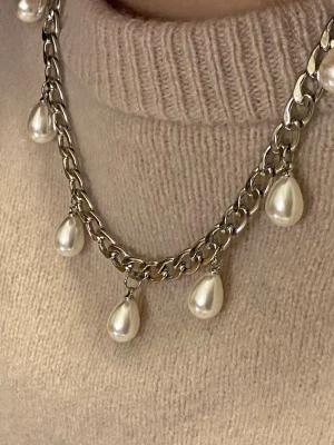 Silverfärgat halsband med pärldroppar - Chunky halsband i silverfärgad rostfritt stål med sju hängande droppformade pärlor. Kedjan är grov och ger ett trendigt intryck, medan pärlorna ger en elegant touch. Perfekt för att lyfta din outfit med en cool och stilren detalj.