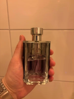 Prada L'Homme Eau de Toilette - Säljer en lyxig parfym från Prada, L'Homme. Flaskan är stilren i genomskinligt glas med silverfärgad metallkork och svart detalj på sidan. Innehållet är lila. Perfekt för dig som gillar exklusiva dofter och snygg design. Ca 65% kvar.