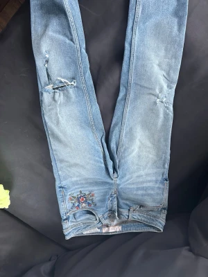 Ljusblå jeans med broderi - Säljer ett par ljusblå jeans med slitna detaljer och råa benslut. Jeansen har ett färgglatt blommigt broderi vid ena fickan framtill. Modellen är smal och materialet är klassisk denim. Perfekta för dig som gillar en avslappnad och trendig stil.