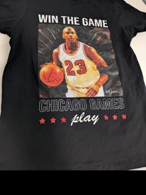 Svart t-shirt Chicago Games - Cool svart t-shirt från Celebrity Icons med stort tryck av basketspelare och texten 'WIN THE GAME' samt 'CHICAGO GAMES play' med röda stjärnor. Perfekt för dig som gillar sport och streetstyle. Rund halsringning och korta ärmar.