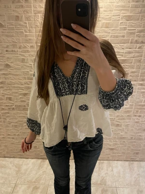 Vit boho-blus med svart brodyr Zara - Luftig vit boho-blus från Zara med svart brodyr och vida ärmar. V-ringad hals med dekorativ knytning och tofsar. Blusen har svarta broderade detaljer på ärmslut, halsringning och spridda mönster över hela plagget. Perfekt till jeans eller kjol.