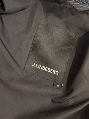 Svart jacka från J.Lindeberg - Stilren svart jacka från J.Lindeberg med hög krage, dragkedja och knappar framtill. Jackan har flera fickor med lock och knappdetaljer samt lång ärm. Perfekt för dig som vill ha en klassisk look med modern touch.