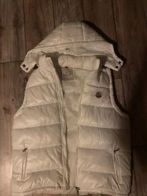 Vit väst från Moncler med huva - Snygg vit dunväst från Moncler med avtagbar huva och dragkedjor på fickorna. Klassisk Moncler-logga på bröstet och quiltad design. Perfekt för lager-på-lager och streetstyle. Materialet är glansigt och fyllt med dun för extra värme.