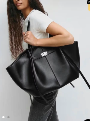 Svart shoppingväska i skinnimitation - Stilren svart shoppingväska i skinnimitation med silverfärgat spänne och långa handtag. Väskan har en rymlig design och dekorativa knytband på sidorna. Perfekt för dig som vill ha plats för allt du behöver under dagen.