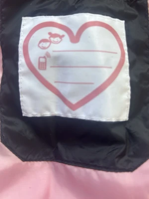 Vårjacka - Svart pufferjacka med rosa foder - Snygg svart pufferjacka med ljusrosa foder och huva. Jackan har dragkedja framtill och två fickor med dragkedja. Insidan har en söt etikett med hjärtmotiv. 