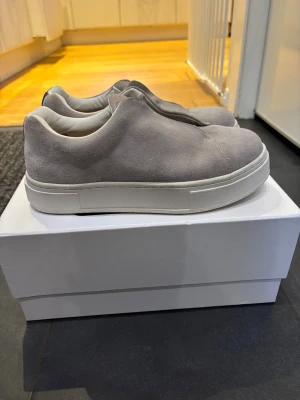 Gråa slip-on sneakers i mocka - Stilrena gråa slip-on sneakers i mjuk mocka med vit, chunky sula. Skorna har minimalistisk design utan snörning och en clean look som passar till många outfits. Insidan är fodrad i vitt för extra komfort.                                                                         Nästan aldrig använda så inga skavanker eller liknande. 