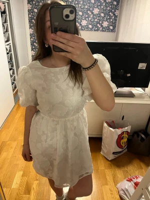 Vit kort studentklänning med puffärmar - Säljer en vit kort studentklänning med puffiga kortärmar och subtilt blommigt mönster. Klänningen har rund halsringning och markerad midja för en feminin siluett. Perfekt för sommarens alla tillfällen! Helt ny!