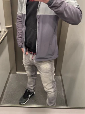 Grå vindjacka från Under Armour - Snygg tvåfärgad vindjacka från Under Armour i ljus- och mörkgrått. Jackan har dragkedja, huva och elastiska muddar vid ärmslut. Perfekt för blåsiga dagar och sportiga outfits. Under Armour-logga på bröstet ger en sportig vibe.