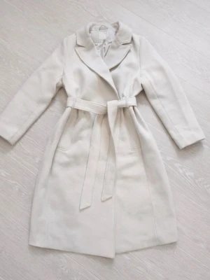 Beige kappa med knytskärp från H&M - Stilren beige kappa från H&M med klassisk krage och knytskärp i midjan. Kappan har två fickor framtill och är helfodrad för extra komfort. Perfekt för dig som vill ha en enkel men elegant look till hösten eller vintern. Säljer den pga den inte kommer till användning. Använd fåtal gånger. 