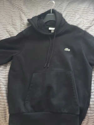 Svart hoodie från Lacoste - Snygg svart hoodie från Lacoste med klassisk krokodillogga på bröstet. Hoodien har stor känguruficka framtill, dragsko i huvan och ribbade muddar. Tillverkad i mjukt bomullsmaterial som känns skönt mot huden. Perfekt för en avslappnad och stilren look.