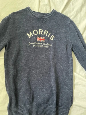 Blå stickad tröja från Morris - Mörkblå stickad tröja från Morris med broderad text och brittisk flagga på bröstet. Tröjan har rund halsringning, ribbade muddar och är tillverkad i mjukt material. Perfekt för dig som gillar klassisk och stilren design.