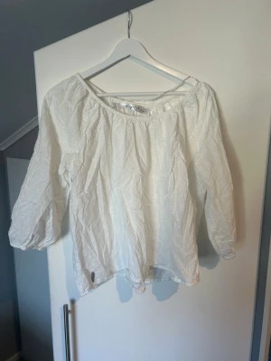 Vit broderad blus med trekvartsärm - Säljer en vit blus med broderade detaljer och trekvartsärmar. Blusen har en offshoulder-design med resår runt halsen och är tillverkad i ett lätt och luftigt material. Perfekt för en avslappnad och trendig look.