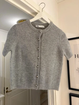Grå kortärmad kofta från Zara - Säljer en ljusgrå, kortärmad kofta från Zara med rund halsringning och silvriga knappar framtill. Koftan är stickad i mjukt material och har ribbade kanter vid ärmslut och nederkant. Perfekt att styla över en topp eller klänning.