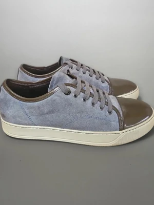 Lanvin  - Snygga sneakers i grå mocka med bruna detaljer i läder på tå och häl. Skorna har vita sulor och matchande grå snörning. Perfekta för dig som gillar en stilren och unik look med mix av material.