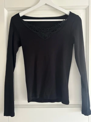 Svart långärmad topp med spets - Snygg svart långärmad topp med v-ringad hals och dekorativ spetsdetalj framtill. Materialet känns mjukt och stretchigt, vilket gör den bekväm att bära hela dagen. Köpt från Zalando - märke only. Helt oanvänd!