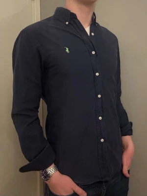 Ralph Lauren skjorta – M slim fit - Säljer denna snygga blå Ralph Lauren skjorta med grön logga i storlek M, Slim Fit. Snygg och stilren modell som passar till både vardag och finare tillfällen. I bra men använt skick.  Om du har några frågor tveka inte att höra av dig. Allt gott Deluxe Garderoben. 