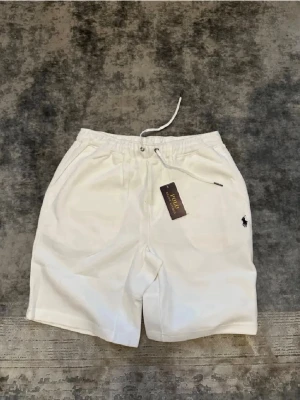 Vita shorts från Polo Ralph Lauren - Fräscha vita shorts från Polo Ralph Lauren med elastisk midja och snörning. Klassisk design med sidofickor och en bakficka. Perfekta för sommaren och har den ikoniska loggan broderad på ena benet.