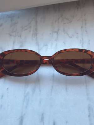 Bruna ovala solglasögon leopardmönster - Snygga ovala solglasögon med brunt leopardmönstrat båge och bruna glas. Perfekt accessoar för att lyfta din stil med en retrovibe. Passar dig som vill sticka ut och addera lite edge till din look.