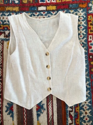 Beige linneväst med träknappar - Stilren beige väst i linne med fyra träknappar framtill och v-ringad hals. Västen har en rak passform och är ärmlös, perfekt för en avslappnad men ändå trendig look. Materialet är svalt och luftigt linne.