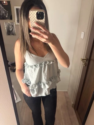 Ljusblå volangtop med v-ringning - Säljer en ljusblå linnetopp med dubbla volangdetaljer och v-ringad hals. Toppen är ärmlös och har en lös passform som ger en avslappnad och trendig look. Perfekt att styla med jeans eller kjol för en fräsch outfit.