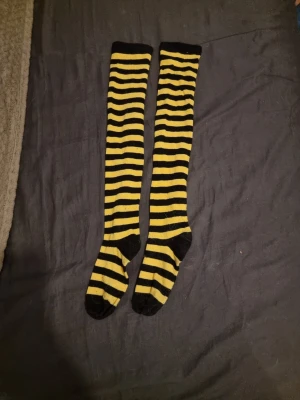 Randiga knästrumpor i svart och gult - Säljer ett par långa knästrumpor med svart och gul randigt mönster. Strumporna har svarta tår, hälar och muddar. Perfekta för att sticka ut med en färgglad och lekfull stil.