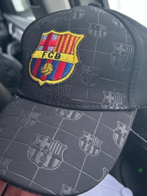 Svart FC Barcelona keps - Snygg svart keps med FC Barcelona-logga broderad framtill och diskret mönster av klubbens emblem över hela kepsen. Justerbar baktill och tillverkad i slitstarkt tyg. Perfekt för dig som vill representera ditt favoritlag med stil. Skriv gärna för frågor!