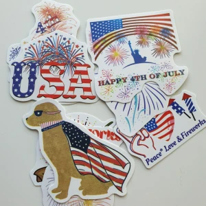 9st Klistermärken med USA-tema och fyrverkerier - Ett set med färgglada klistermärken med USA-tema, fyrverkerier, flaggor och texter som 'Happy 4th of July' och 'Peace Love & Fireworks'. Motiven inkluderar hund med amerikansk flagga, frihetsgudinnan och patriotiska symboler. Perfekt för dekoration eller scrapbooking.