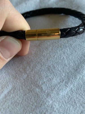 Brunt flätat läderarmband - Säljer ett stilrent brunt armband i flätat läder med metallspänne. Armbandet har en rund form och passar perfekt för dig som gillar klassiska accessoarer med en modern touch. Perfekt att kombinera med andra smycken för en personlig look.