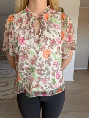 Blommig blus med knytning - Söt blus i tunnt material med blommigt och mönster i rosa, orange, grönt och brunt. Knytning och liten volang vid halsen samt korta, luftiga ärmar. Knappt använd 