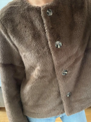 Brun pälsjacka med knappar - Säljer en brun pälsjacka med stora, dekorativa knappar framtill. Jackan har rak passform, rund halsringning och långa ärmar. Perfekt för dig som vill ha en mjuk och trendig look under kalla dagar.