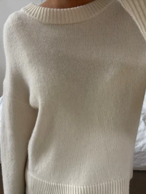 Krämvit stickad tröja i ullmix - Mysig krämvit stickad tröja med rund halsringning och ribbade muddar. Tröjan har en enkel och stilren design, perfekt för lager-på-lager och passar till många olika outfits. Materialet känns mjukt och värmande, troligtvis en ull- eller kashmirblandning.