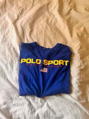 Blå Polo Sport t-shirt från Ralph Lauren - Säljer en blå Polo Sport t-shirt från Ralph Lauren med stor gul logga och amerikansk flagga på bröstet. Klassisk passform och rund hals. Perfekt för dig som gillar sportig och clean stil.