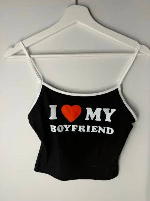 Svart linnetopp med text och hjärta - Cool svart linnetopp med vita kanter och tunna axelband. Framsidan har tryck med texten 'I ❤️ MY BOYFRIEND' i vitt och rött. Perfekt för att visa lite attityd och kärlek. Materialet känns mjukt och stretchigt.