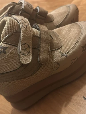 Beige sneakers med kardborre och mönster - Snygga beige sneakers med högt skaft och kardborreband. Skorna har ett unikt blommigt mönster och är gjorda i en mix av textil och mockaliknande material. Sulan är tjock och ger en cool chunky look. Perfekta för dig som vill sticka ut med din stil.