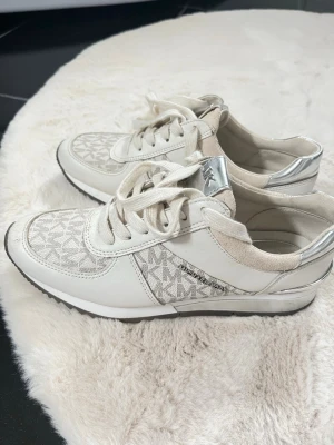 Vita sneakers från Michael Kors - Snygga vita sneakers från Michael Kors med silverdetaljer och logomönster på sidan. Skorna har låga skaft, snörning och en mix av textil och lädermaterial. Perfekta för dig som gillar stilrena och trendiga skor.size 36/37